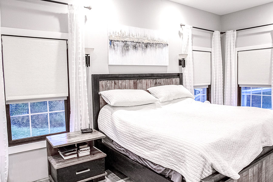 Find the perfect style and color of window shade for any room and then ask to have it motorized! White Roman shades on windows in a farmhouse-style bedroom
