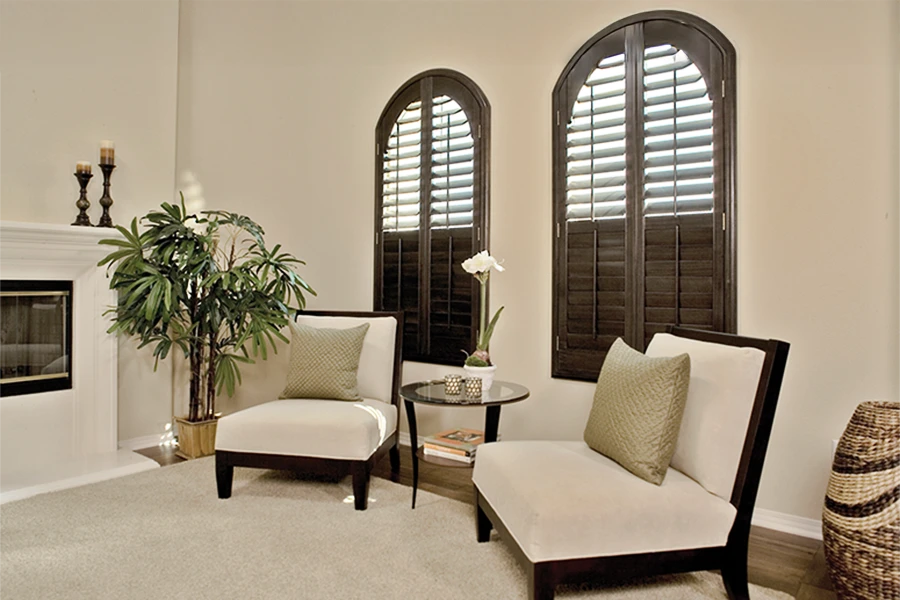 Whatever the cost, interior shutters are the perfect choice for specialty-shaped windows. Dark wood shutters on two arched windows in a living room